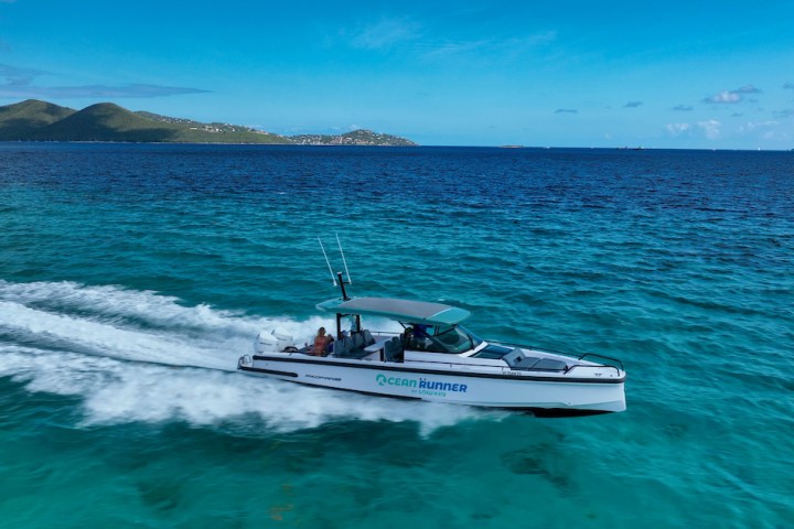 Speedboat cruising on clear blue ocean with distant hills under a bright blue sky.