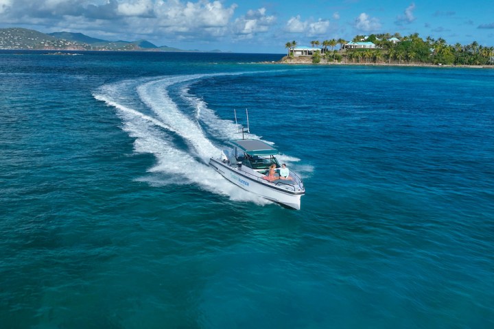 A speedboat cruising near a tropical island with clear blue waters and a cloudy sky.