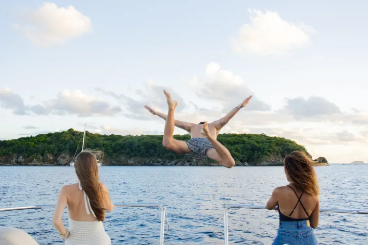 Person jumping off boat into water, two others watching, island in background.
