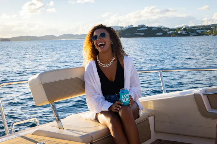 Woman laughing on a boat, holding a can, wearing sunglasses and a white shirt.