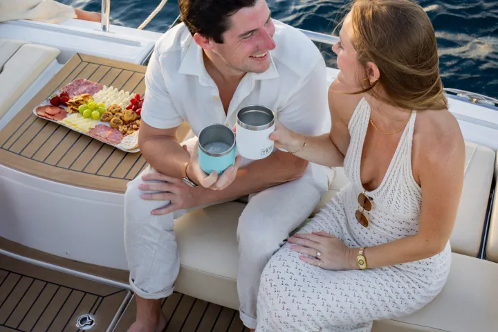 Couple sitting on a boat, toasting with cups, charcuterie board nearby.
