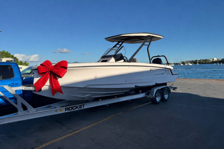 White boat with red bow on trailer by blue truck near water under blue sky.