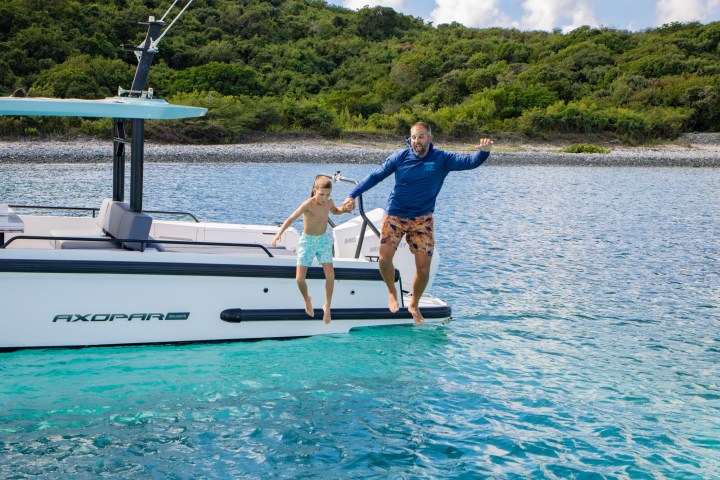 Two people jumping off a boat into clear blue water near a forested shoreline.