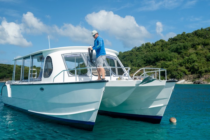 Person standing on a white catamaran boat on blue water, with a lush green island in the background.