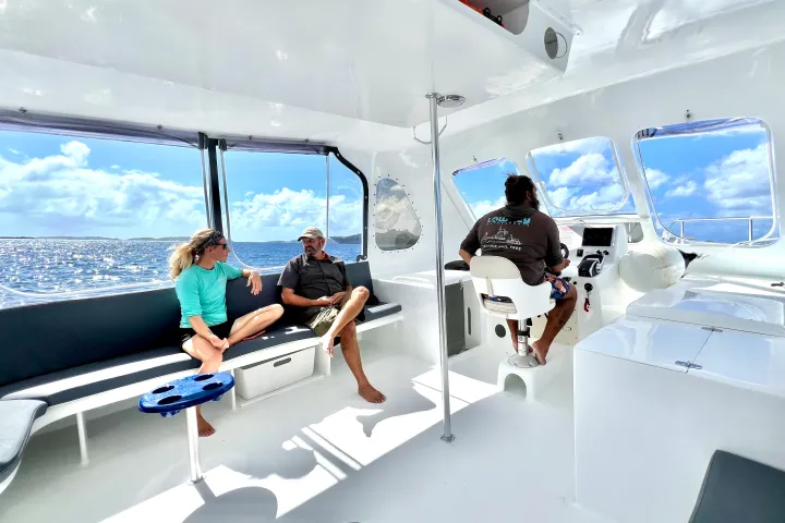 Three people on a boat; two seated on bench talking, one steering. Bright interior with ocean view.